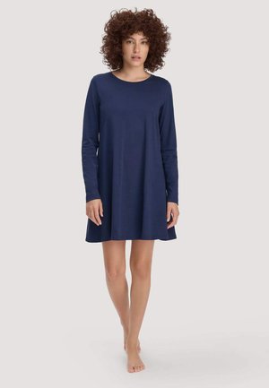 Frau mit lockigem Haar, die ein langärmliges, knielanges marineblaues Kleid trägt und barfuß vor einem schlichten hellen Hintergrund steht.