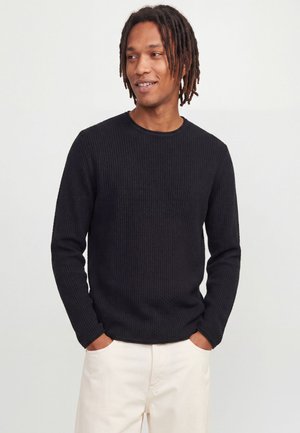 Homme avec des dreadlocks portant un pull noir côtelé à manches longues et un pantalon blanc, mains dans les poches, regardant sur le côté avec un léger sourire.