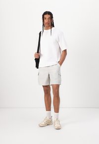 Camiseta blanca de gran tamaño, pantalones cortos de cargo gris claro con bolsillos, zapatillas beige con detalles texturizados, de pie frente a un fondo sencillo.