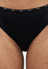 Sous-vêtements noirs avec le logo blanc répétitif "BOSS" sur la ceinture, portés sur des hanches et des cuisses supérieures couleur peau.