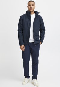 Mørkeblå softshell-jakke med hette, glidelås og frontlommer, kombinert med en hvit t-skjorte med rund hals og mørkeblå bukser, båret med joggesko.