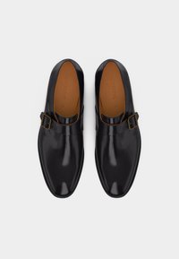 Paire de chaussures monk en cuir noir brillant avec boucle dorée, vues de dessus sur un fond clair uni.