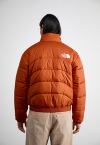Person som bär en rostfärgad puffjacka med The North Face-logotyp på ryggen, i kombination med beige byxor, stående mot en enhetlig bakgrund.