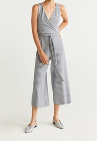 Combinaison rayée grise avec un décolleté en V profond, un design portefeuille, des jambes larges et raccourcies, ainsi qu'une ceinture assortie. Portée avec des chaussures grisées à enfiler.