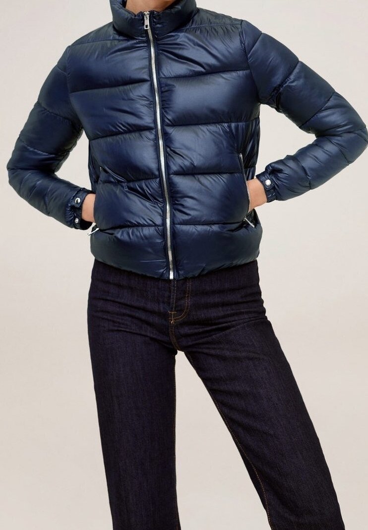 Veste matelassée bleu marine avec matelassage horizontal, col montant et fermeture éclair. Les manches sont effilées avec des poignets élastiques. Portée avec un jean foncé.