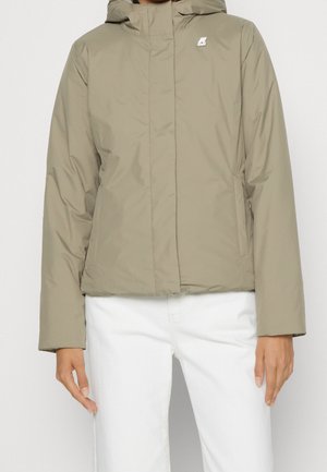 Woman wearing a light brown hooded puffer jacket over white pants, standing with arms relaxed at sides against a plain background.