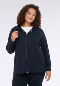 Felpa con zip navy in morbido tessuto, dotata di cappuccio, zip color oro e dettagli laterali. Vestibilità comoda con maniche lunghe.