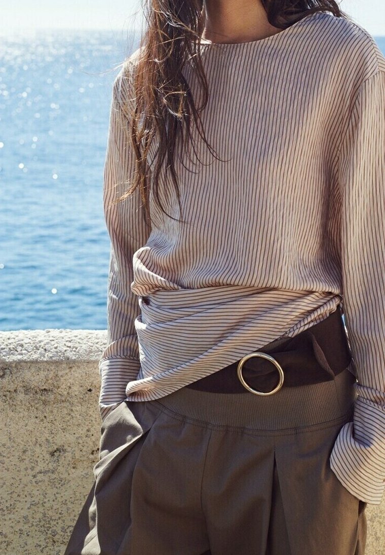Femme portant une chemise légère à manches longues rayée rentrée dans un pantalon marron taille haute avec une large ceinture, debout près d'une rambarde en pierre près de l'eau.