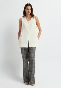 Top de punto sin mangas color crema con escote en V y botones en la parte delantera. Combinado con pantalones de pierna ancha de cuadro gris y zapatos de tacón beige. Textura suave.
