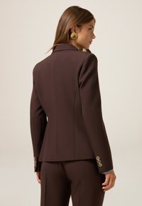 Blazer marrón ajustado con un ajuste estructurado, cuello afilado, cintura entallada y puños abotonados. Tejido suave con un acabado limpio.