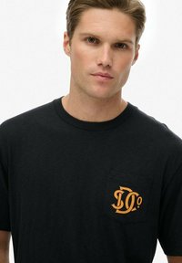 Camiseta de algodón negra con un bolsillo en el pecho izquierdo y un diseño gráfico naranja con letras. Cuello redondo clásico y mangas cortas.