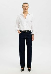 Camisa blanca de botones con mangas largas, combinada con jeans de pierna recta azul oscuro y zapatos negros de punta. Diseño simple y clásico.