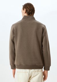 Brauner Pullover-Jacke mit hohem Kragen, gerippten Bündchen und Saum. Glatte Textur mit lockerer Passform, von hinten gezeigt.