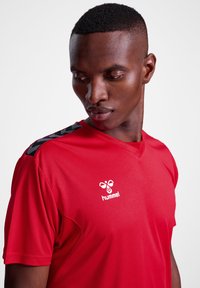 Camiseta deportiva roja de manga corta, con cuello en V, que presenta detalles negros en los hombros y un logo blanco en el pecho. Textura de tela suave.