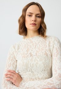 Pull en tricot blanc avec un motif florale en dentelle, des manches longues et un col haut avec une texture côtelée subtile.