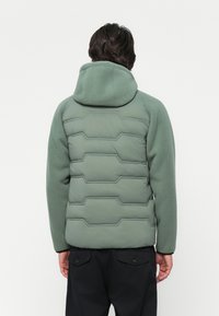 Veste rembourrée verte avec un dos texturé et matelassé, dotée d'une capuche et d'accents noirs, associée à un pantalon sombre. Matériau doux et chaud.