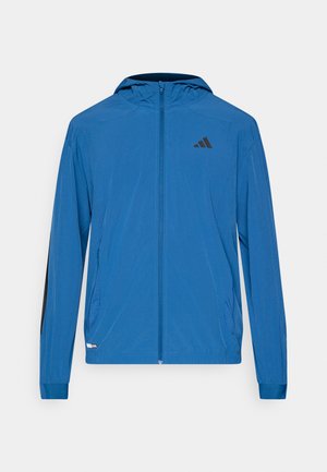 Veste légère bleu avec fermeture éclair et capuche, avec des accents noirs sur les manches et un logo noir sur la poitrine en haut à gauche.