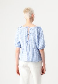 Blå og hvit rutete bluse med pufferm, knyttet bak på nakken, og peplum-fal den, laget av lett bomullsstoff.