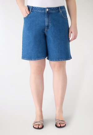 Persona che indossa pantaloncini di jeans blu di media lunghezza con una maglietta bianca e sandali neri a fasce, in piedi contro uno sfondo chiaro e uniforme.