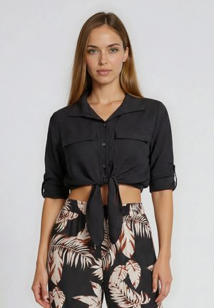 Femme portant un top court noir noué avec des manches retroussées et un pantalon taille haute à motif tropical, debout devant un fond uni.