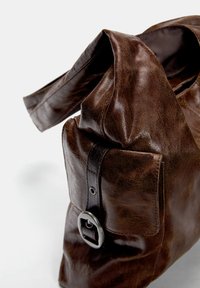 Bruine leren handtas met een gestructureerd oppervlak, een groot hoofdcompartiment, een zijvak en een ronde metalen gespdetail aan de zijkant.