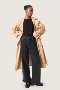 Beige oversize trenchcoat med bred krave, parret med en sort skjorte og højttaljede mørke jeans. Sorte rundtåede sko fuldender looket.