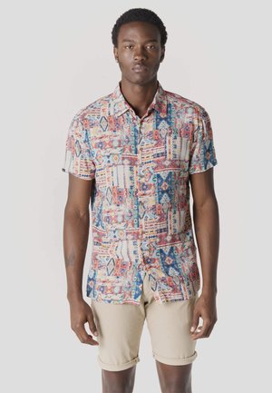 Joven con una camisa de manga corta de estampado colorido y pantalones cortos beige remangados, de pie contra un fondo liso.