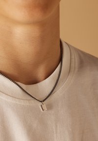 Zilveren rechthoekige hanger aan een donkere ketting, gedragen over een wit T-shirt met ronde hals. De hanger hangt net onder de kraag.