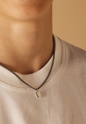 Zilveren rechthoekige hanger aan een donkere ketting, gedragen over een wit T-shirt met ronde hals. De hanger hangt net onder de kraag.