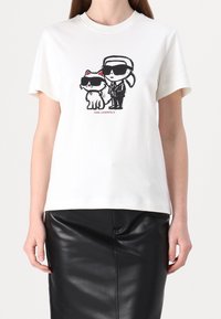 Bílý bavlněný tričko s grafikou postavy a kočky, obě ve stylových černých slunečních brýlích, a textem "KARL LAGERFELD" pod nimi.
