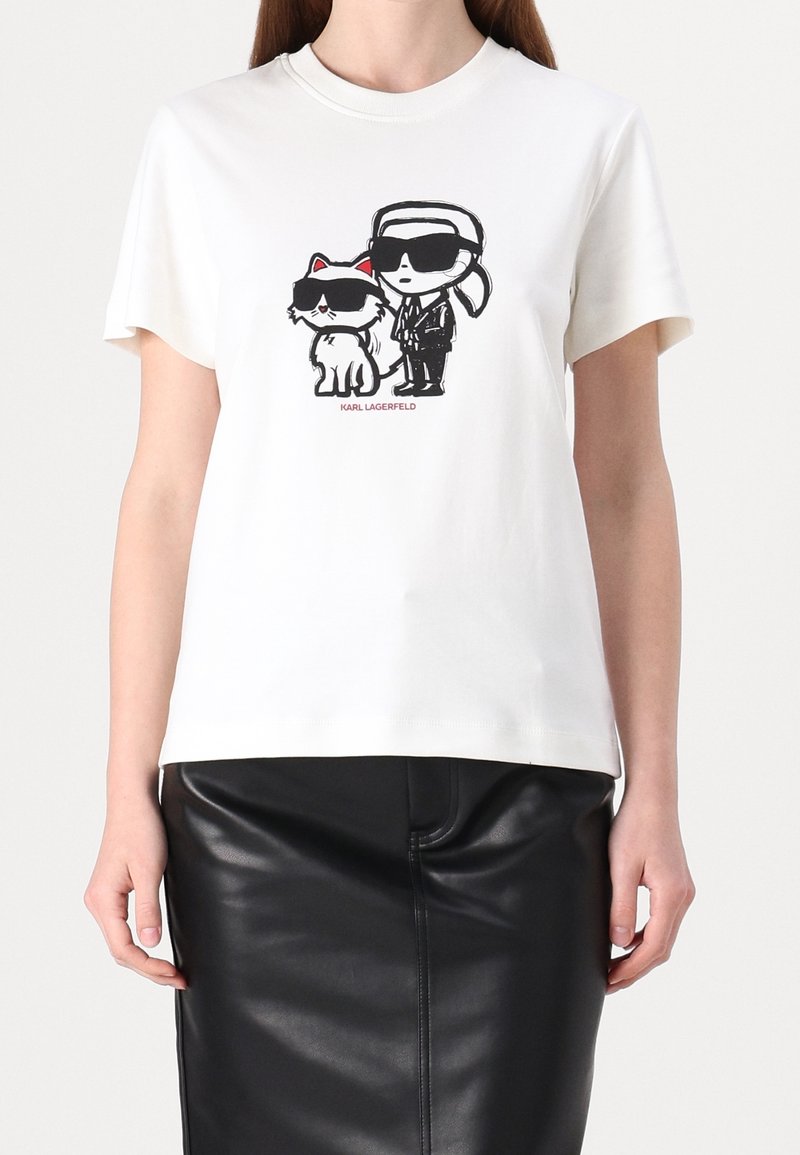 Bílý bavlněný tričko s grafikou postavy a kočky, obě ve stylových černých slunečních brýlích, a textem "KARL LAGERFELD" pod nimi.