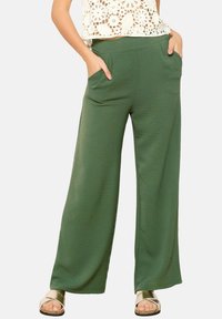Wide-leg green pants made of textured fabric, featuring side pockets and a high waist. Paired with flat sandals.