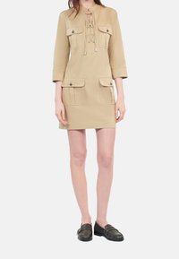 Woman wearing beige lace-up pocketed dress with three-quarter sleeves and black slip-on loafers against white background.