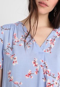 Vestido floral de color azul claro en tela suave, con escote en V, mangas cortas y patrones de flores blancas y rosas sobre ramas.