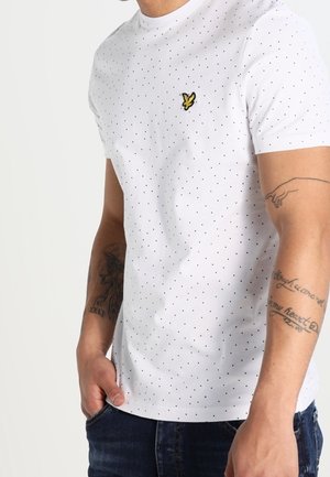 Biała koszulka z krótkim rękawem z małymi czarnymi kropkami i żółtym logo ptaka na piersi, noszona przez osobę z tatuażami, z ręką w kieszeni.