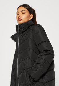 Czarna kurtka puchowa z wysokim kołnierzem, zapięciem na zamek błyskawiczny i przeszyciem w wzór w zygzak. Posiada boczne kieszenie oraz gładką, lekką fakturę.