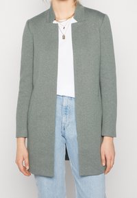 Manteau gris clair long avec un col structuré et sans fermetures. Doté de manches longues et d'un design à l'avant ouvert. Porté par-dessus un haut blanc.