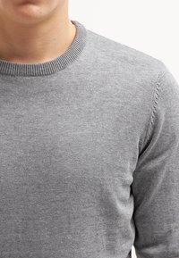 Nahaufnahme einer Person, die einen schlichten grauen Strickpullover mit Rundhalsausschnitt trägt, wobei der Hals und der obere Oberkörper vor einem weißen Hintergrund zu sehen sind.