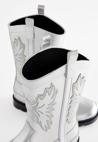 Silberne Gummistiefel mit glänzender Oberfläche, schwarzer Innenverkleidung, erhabenen Blattmustern, kontrastierenden Nähten und Laschen am oberen Rand.
