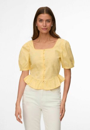 Kobieta z długimi brązowymi włosami, ubrana w żółtą bluzkę z bufkami na rękawach i guzikami z przodu oraz białe spodnie, na jednolitym tle.