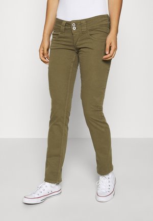 Personne portant un pantalon slim fit vert olive, des baskets blanches et un haut blanc, debout devant un fond clair uni.