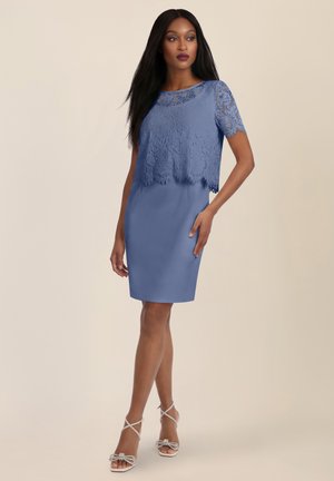 Frau, die ein knielanges blaues Spitzenkleid mit kurzen Ärmeln trägt, steht mit einer Hand in der Hüfte und weißen Riemchen-High Heels.