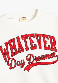 Witte sweatshirt met rode bedrukte tekst "WHATEVER Day Dreamer," met een versleten effect. Ribbelkraag en een zachte stofstructuur.