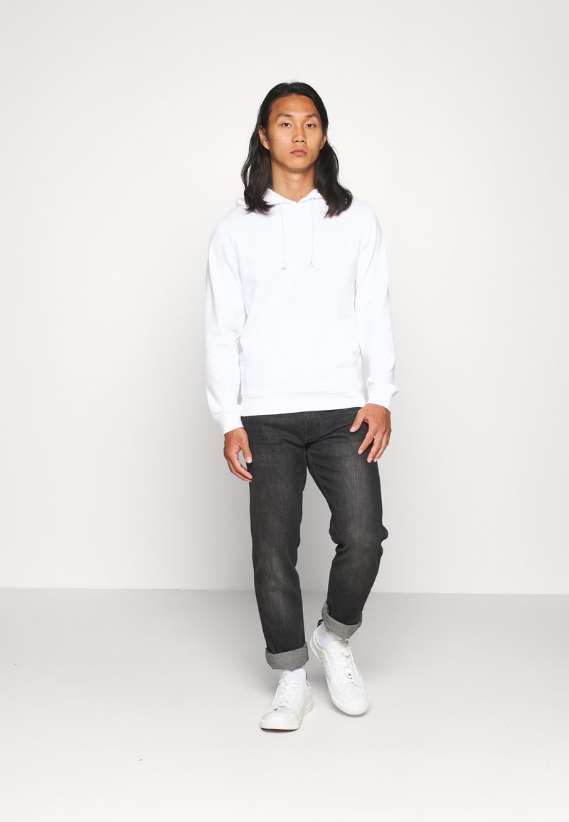 Haut à capuche blanc avec cordon de serrage et poches latérales, associé à un jean délavé foncé. Le mannequin porte des baskets blanches, debout sur un fond clair.