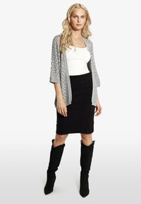 Stickad grå öppen cardigan över vit topp, kombinerad med en svart pennkjol och knähöga svarta stövlar med en strukturerad design.