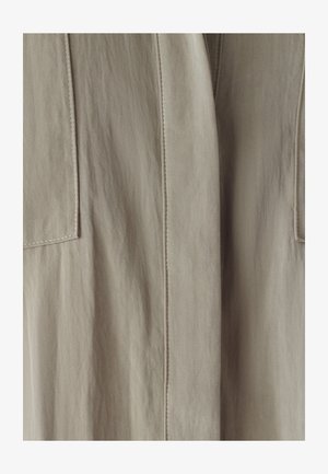 Beige stof met een gladde textuur, met opvallende verticale naden en twee vierkante zakken aan elke kant.