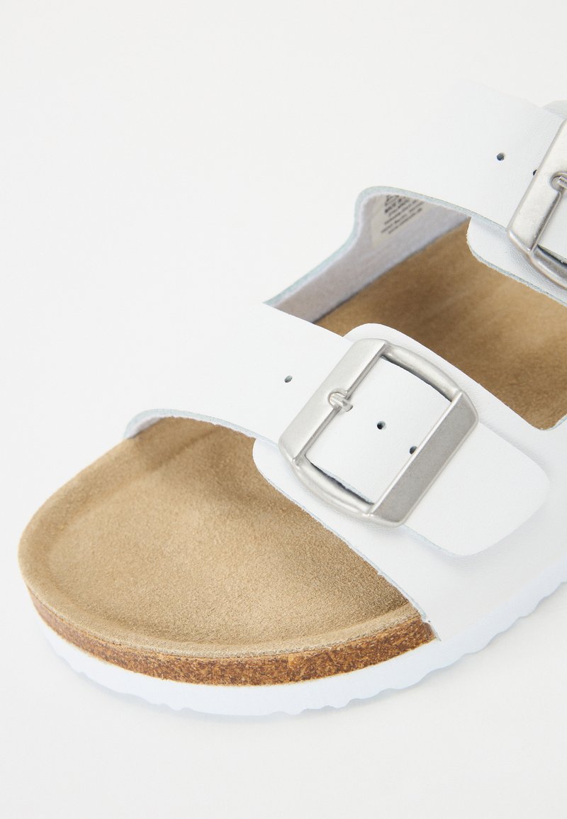 Sandale blanche avec deux sangles réglables, boucles en argent, et une semelle intérieure en brun texturé. Caractérisée par une semelle en caoutchouc léger avec un bord en liège.