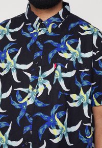 Chemise noire à manches courtes avec un motif floral bleu et jaune, design boutonné et une poche poitrine avec un détail d'étiquette rouge.