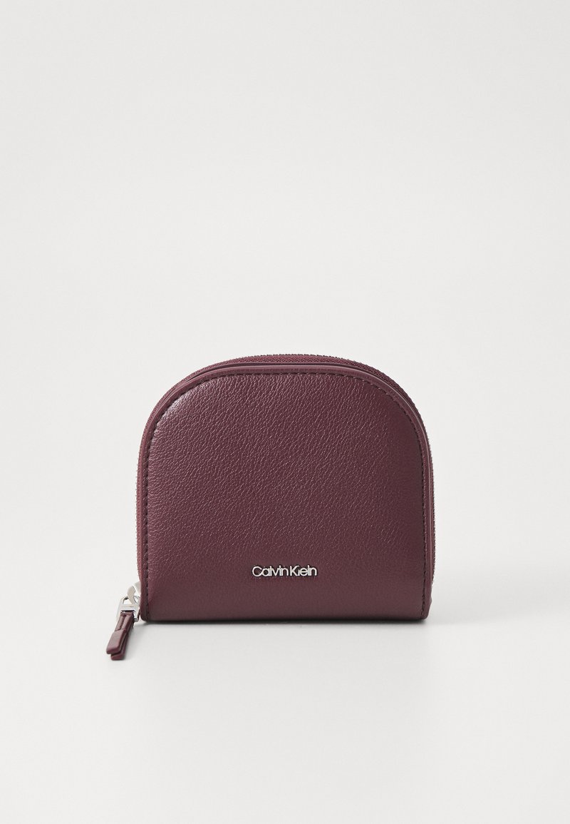 Bordowy skórzany portfel o zaokrąglonym kształcie, o błyszczącej powierzchni, zamykany na zamek błyskawiczny, z srebrnym logo Calvin Klein z przodu.