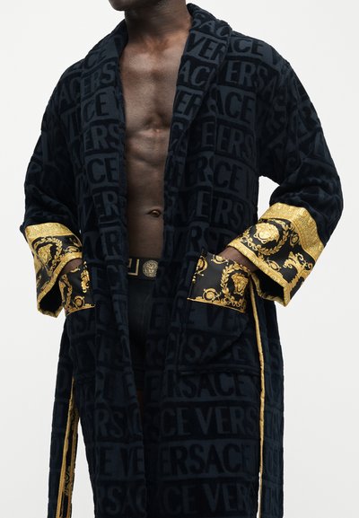 Μαύρο ρόμπα με ανάγλυφο κείμενο "VERSACE", μανσέτες και τσέπες με χρυσό μοτίβο, φορεμένο ανοιχτό πάνω από γυμνό στήθος και μαύρα σορτς με πόρπη ζώνης Medusa.